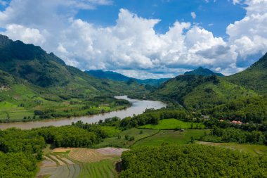Dolambaçlı bir nehir, kuzey Laos 'taki dramatik kümülüs bulutlarının altında yemyeşil tepeleri ve katmanlı pirinç teraslarını keser. Manzara yoğun tropikal bitki örtüsü, yuvarlanan dağlar ve canlı yaz ışığına sahiptir..