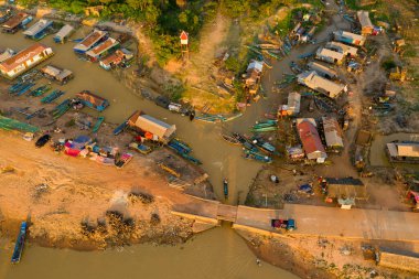 Kamboçya 'daki Tonle Sap yakınlarındaki çamurlu kıyılarda yüzen evleri, renkli tekneleri ve derme çatma sığınakları olan nehir kenarındaki bir köye ait hava perspektifi. Sahne sıcak akşam güneşiyle yıkanır, eşsiz yerleşim yapısını vurgular ve...
