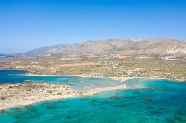 Yunanistan 'ın Girit kentindeki Elafonisi plajında sığ bir turkuaz körfez, kumlu tükürük ve canlı mavi gökyüzünün altındaki kurak dağlar görülüyor. Sahne parlak ve geniş, Akdeniz açıklığını çağrıştırıyor..