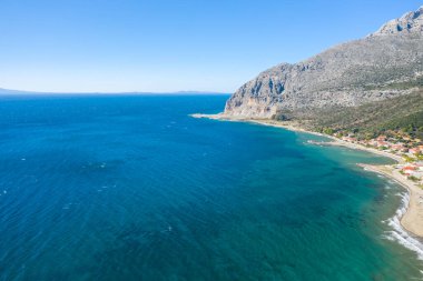 Dramatik bir kayalık dağ ve Yunanistan 'ın batısındaki küçük bir sahil köyüyle çevrili geniş bir turkuaz kıyı şeridinin havadan görünüşü. Açık gökyüzü ve güneş ışığı dalgalı denizi ve desenli kayalıkları aydınlatır..