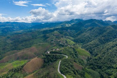 Laos 'un Phonsavan ve Phou Khoun arasındaki bölgede yemyeşil tepelerden ve dağınık kırsal evlerden geçen eğimli bir dağ yolunun havadan görünüşü. Geniş arazide yuvarlanan arazi, yoğun bitki örtüsü ve altında dramatik bulutlar yer alır.