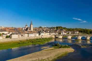 Parlak güneş ışığı ön planda Loire nehri ve yemyeşil yeşillik ile La Charite sur Loire 'ın tarihi manastır kulesi ve klasik taş köprüsünü vurgular. Sahne, ortaçağ mimarisinin ahenkli bir harmanını yakalıyor.