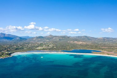 Sardinya 'daki Spiaggia Lu Impostu' nun geniş hava manzarası, canlı turkuaz sular, beyaz kumlu plaj ve kayan tepeler ve dağınık bulutlar üzerine kurulu bir lagün sergileniyor..