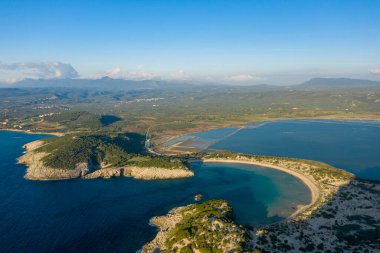 Yunanistan 'ın Messinia kentindeki ikonik yarı dairesel Voidokilia Sahili' nin turkuvaz suları, altın kumu ve engebeli burunlar ve kayan tepelerle çevrili bir kıyı lagünü manzarası. Açık mavi gökyüzü ve geniş Akdeniz manzarası oluşturur