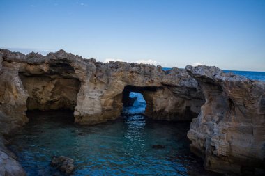 Doğal taş kemer oluşumları, İtalya 'nın Marina Serra kentinin engebeli kıyı şeridi boyunca berrak turkuaz suyun üzerinde yükselir. Sahnede kireçtaşı kayalıklar ve açık bir gökyüzünün altında sakin mavi bir deniz yer alıyor..