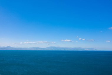 Geniş açık deniz manzarası derin mavi su ve ufukta soluk bir dağ silsilesi hepsi bulutlu, canlı bir gökyüzünün altında. Sahne, ferah ve sakin bir şekilde, dingin bir gün ışığı eşliğinde, dingin atmosferi canlandırır..