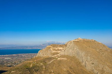 Antik taş kalıntılarıyla taçlandırılmış engebeli Akrokorinth tepesinin insansız hava aracı görüntüsü, Yunanistan 'da parlak ve berrak bir gökyüzünün altında parıldayan mavi uçuruma ve yayılan ovalara tepeden bakıyor.