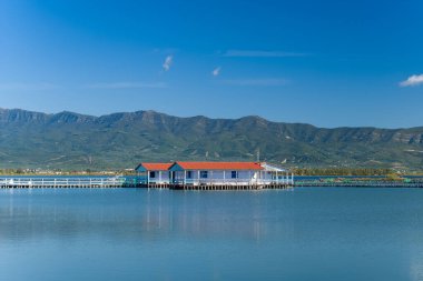 Yunanistan 'ın batısındaki Missolonghi tuz gölünün sakin sularının üzerinde kırmızı çatılı geleneksel stilt evi bulunuyor. Sahnede berrak mavi gökyüzü, uzak yeşil dağlar ve huzurlu doğal ortamdaki yansımalar yer alıyor..
