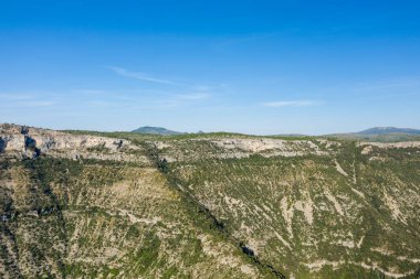 Dik uçurumlar, dağınık yeşil bitki örtüsü ve dramatik manzaranın üzerinde uzanan berrak mavi gökyüzü olan Navacelles Sirki 'ndeki engebeli kireçtaşı kanyonunun geniş hava perspektifi..