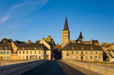 Altın güneş ışığı, şehrin girişindeki taş köprüden görülen La Charite sur Loire 'ın tarihi manastır kulesini ve kümelenmiş çatı katlarını aydınlatır. Açık mavi gökyüzü ve boş cadde huzur dolu bir Loire Vadisi sabahını anımsatıyor..