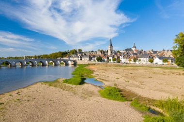 Fransa 'nın merkezindeki La Charite sur Loire' a geniş bir manzara, Loire Nehri üzerinde tarihi bir taş köprü, yeşil bantlı kumlu nehir kıyısı ve dinamik bir gökyüzü altında geleneksel çatıların üzerinde yükselen kilise kulesi. Sahne vurgulanıyor