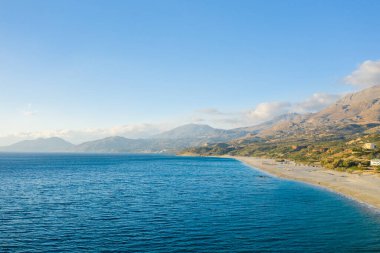 Kıvrımlı kumlu kıyı şeridi Girit 'teki Triopetra Plajı' nda güneşli engebeli dağlarla ve açık bir gökyüzünün altında dağınık yeşilliklerle buluşuyor. Huzurlu manzara açıklık ve doğal güzellik yayar..