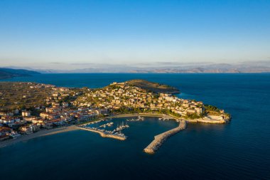 Bir marinaya ait Paralio Astros 'un hava manzarası, yarımadada kümelenmiş evler, ve berrak akşam gökyüzünün altında geniş mavi deniz. Huzurlu kıyı manzarası, ufuktaki uzak dağlarla sıcak güneş ışığıyla yıkanır..