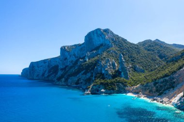 Kireçtaşı uçurumları canlı turkuaz suların ve Cala Goloritze, Baunei, Sardinya yakınlarında tenha bir kıyı şeridinin üzerinde yükselir. Güneşli Akdeniz manzarası açık mavi gökyüzü altında yemyeşil ve dramatik kıyı oluşumlarına sahiptir..