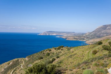 Yunanistan 'ın Vathia kenti yakınlarındaki açık mavi bir kıyı şeridine inen yuvarlanan yeşil tepelerin geniş manzarası. Uzak dağlar ve sakin Akdeniz sularıyla berrak bir gökyüzü altında güneşli bir manzara..