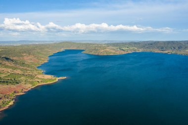 Lac de Salagou 'nun derin mavi su manzarası engebeli yeşil tepeler ve kırmızı toprak ile buluşuyor dağınık bulutlar ve uzak dağlarla dolu bir gökyüzünün altında..