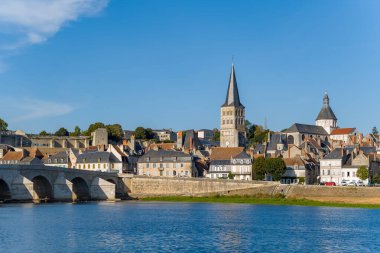 La Charite sur Loire 'nin siluetinde tarihi bir taş köprü, geleneksel evler ve nehir kenti üzerinde yükselen önemli bir kilise kulesi yer almaktadır. Parlak gün ışığı ve açık mavi gökyüzü ortaçağ mimarisi ve sakin Loire nehrinin altını çiziyor.
