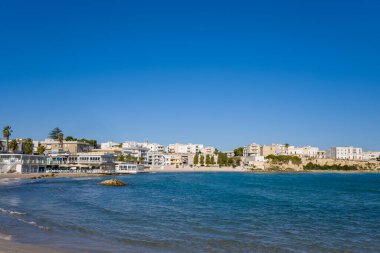 Beyazlatılmış binalar Adriyatik Denizi boyunca uzanan Otranto sahili boyunca uzanırken, hafif dalgalar gün ışığında kumlu kıyılara vuruyor. Sahnede modern kıyı mimarisi, mavi su ve güney İtalya 'da geniş bir canlı gökyüzü yer alıyor..