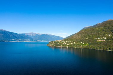 Kuzey İtalya 'daki Maggiore Gölü' nün yukarısındaki yeşil tepelere tünemiş Cannobio 'nun geniş manzarası, derin mavi su ve bulutsuz bir gökyüzünün altındaki uzak dağlık tepeler. Sahne, sakin göl manzarası ve yemyeşil bitki örtüsünü vurguluyor..