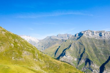 Güneşli çimenli tepelerin geniş manzarası ve Pireneler 'deki Gavarnie Gedre yakınlarında parlak mavi bir gökyüzünün altındaki engebeli kayalık tepeler. Sahnede dramatik bir yükseliş ve açık yaz ışığında canlı doğal dokular yer alıyor..