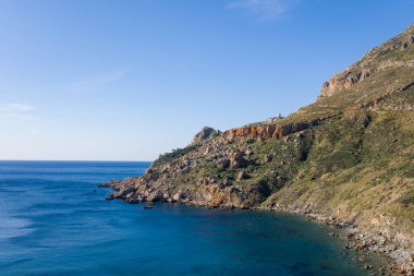 Yunanistan 'ın Cap Tenare kentindeki sakin turkuaz bir körfezin üzerinde yeşil bitki örtüsü parçalarıyla sarp kayalıklar yükseliyor. Temiz güneş ışığı, parlak mavi gökyüzüne karşı engebeli dokuları ve doğal toprak tonlarını ortaya çıkarır..