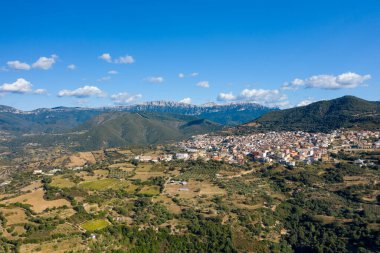 Sardunya 'daki açık mavi gökyüzünün altında, yama alanları ve dramatik dağ zirveleriyle çevrili yuvarlanan tepelere tünemiş Orgosolo' nun hava fotoğrafı..