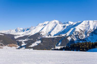 Yoğun çam ormanları ve Fransız Alpleri 'ndeki Megeve yakınlarında açık mavi gökyüzünün altında dramatik dağ tepeleriyle çevrili geniş karla kaplı bir vadi. Kış ışığı, engebeli hatları ve el değmemiş manzarayı vurgular..