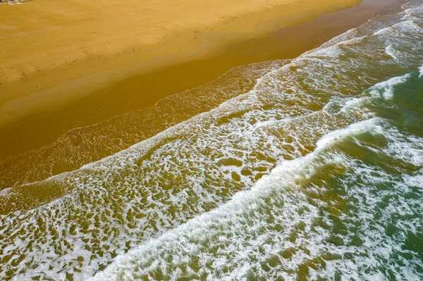 İtalya 'nın Apulia kentindeki Vieste sahilinin geniş altın kumu üzerinde süzülen yumuşak köpüklü dalgaların havadan bakış açısı. Sahne, yumuşak gün ışığında çekilen suyun oluşturduğu dokulu kıyı şeridini ve doğal desenleri vurguluyor..