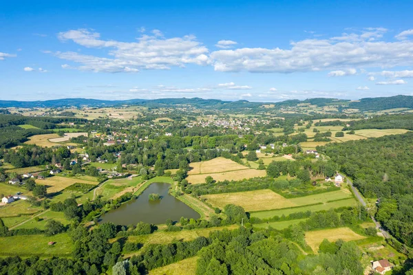Saint-Honore-les-Bains 'in engin hava manzarası. Huzurlu bir gölet, yama alanları ve yemyeşil ve açık mavi bir gökyüzü altında kayan tepelerle çevrili dağınık evler..