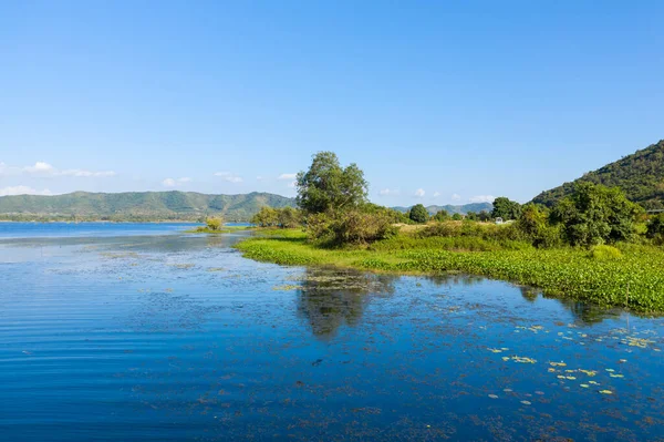 Canlı yeşil bitki örtüsü, sakin mavi suların dağınık ağaçları ve Kamboçya 'nın Kampong Chhnang kentindeki uzak tepeleri yansıttığı Brateak Krola Gölü sınırını çizer. Sahne aydınlık bir gün ışığında berrak bir gökyüzünün altında, doğal bir görüntü sergiliyor.
