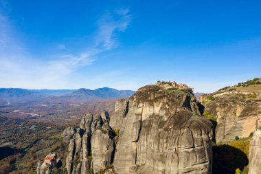 Yunanistan 'ın Meteora kentindeki devasa kumtaşı sütunlara tünemiş tarihi bir manastırın havadan görünüşü, geniş dağ manzaraları ve canlı mavi gökyüzünün altındaki sonbahar ormanları. Dramatik manzarada engebeli uçurumlar ve geniş doğal dokular yer alıyor..