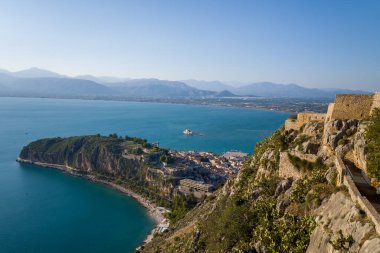 Turkuaz körfezine bakan Akronauplia kalesinin taş surlarından tarihi Nafplio kasabasından ve berrak güneş ışığı altındaki uzak sıradağlardan geniş bir manzara. Kayalık kayalıklar ve antik duvarlar canlı Akdeniz deniz manzarasını çerçeveliyor.