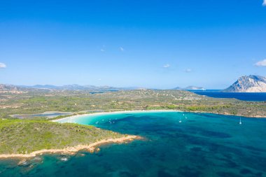 San Teodoro, Sardinya yakınlarındaki Cala Brandinchi sahilinin hava manzarası canlı turkuaz su, demirli yelkenliler, beyaz kumlu bir eğri ve berrak mavi dağların altında yemyeşil bir manzara..