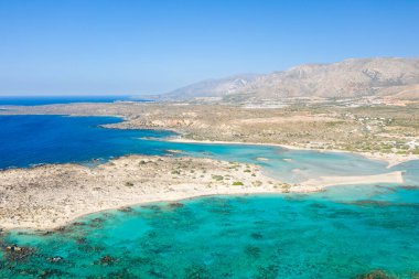 Yunanistan 'ın Girit kentindeki Elafonisi plajının canlı turkuaz koyları, soluk kumları ve berrak mavi gökyüzünün altındaki engebeli dağlarla süpürülen hava manzarası. Manzara güneşli, açık ve doğal olarak davetkar..
