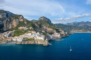 İtalya 'nın Amalfi kentindeki dramatik uçurumların ve beyaz boyalı binaların yakınındaki derin mavi Akdeniz sularında yalnız bir yelkenli sürüklenir. Sahnede engebeli dağlar, teraslı yamaçlar ve gün ışığında ikonik Amalfi Sahili 'nin geniş bir manzarası yer alıyor..