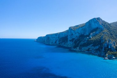 Yoğun yeşil bitki örtüsüyle kaplı yüksek kireçtaşı kayalıkları Cala Goloritze, Baunei, Sardinya yakınlarındaki Akdeniz 'in yoğun mavi sularının üzerinde yükseliyor. Parlak güneş ışığı ve berrak gökyüzü canlı ve sakin bir deniz manzarası yaratır..