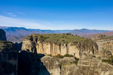 Yunanistan 'ın Meteora kentindeki açık mavi gökyüzü altında yeşil taçlandırılmış geniş kumtaşı kayalıkları, renkli bir sonbahar arkaplanı sağlayan uzak dağlarla çarpıcı bir şekilde yükseliyor..