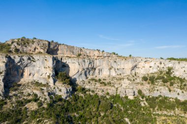 Geniş kireçtaşı kayalıkları, engebeli dokuları ve açık mavi gökyüzü altında alçak bitki örtüsü olan Cirque de Navacelles. Güneş ışığı katmanlı kaya yüzeylerini ve doğal hatları vurgular..