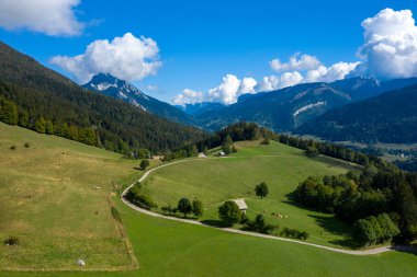 Dolambaçlı bir kır yolu, Chartreuse Massif 'teki yuvarlak yeşil otlakları ve dağınık kırsal evleri, yoğun ormanlar ve canlı mavi gökyüzünün altında dramatik zirvelerle geçer. Sahne bu ikonik ortamın doğal hatlarını ve sakin atmosferini vurguluyor.