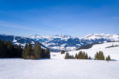 Megeve Alpleri 'nin geniş manzaralı ve açık mavi gökyüzünün altındaki uzak dağ zirveleriyle çevrili engin, karlı kayak yamacı. Parlak kış güneşi canlı dokuları ve huzurlu manzarayı vurgular.