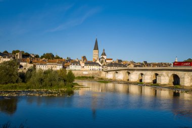 Sıcak akşam güneşi tarihi taş köprüyü, manastır kulesini ve La Charite sur Loire 'nin pastel nehir kenarındaki evlerini yıkıyor. Sakin su ve berrak mavi gökyüzü Loire Vadisi manzarasını güzelleştirir, kasabaların ortaçağ cazibesini ön plana çıkarır..