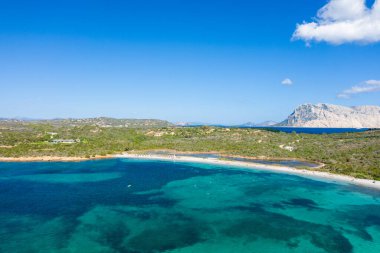 Sardinya 'daki Spiaggia la Salinedda' nın hava görüntüsü, el değmemiş kumlu bir kıyı şeridini, zümrüt mavisi denizi, yemyeşil kıyı bitkisini ve bulutlu bir gökyüzünün altında kayalık bir dağı vurguluyor..