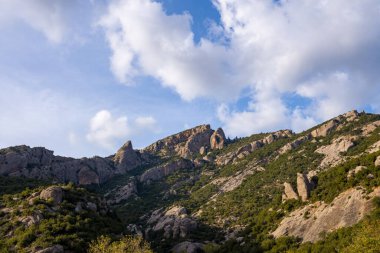 Yeşil çalılar ve dağınık ağaçlarla kaplı yuvarlanan tepelerin üzerinde belirgin kayalık zirveler yükselir, yumuşak bulutlu parlak bir gökyüzüne karşı kurulur. Manzarada engebeli dokular ve açık, güneşli bir çöl hissi var..