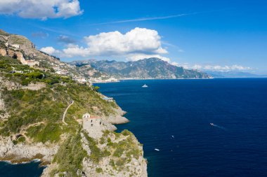 Dik yeşil kayalıklar Amalfi kıyısındaki canlı bir gökyüzünün altında derin mavi Akdeniz sularına iniyor. Dağılmış beyaz bulutlar ve uzak yamaçlardaki köyler bu klasik İtalyan deniz manzarasını tamamlıyor..