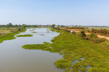 A wide water channel bordered by floating aquatic plants and grassy banks stretches through the countryside near Phnom Penh, Cambodia. A reddish dirt road runs alongside the wetland, with scattered houses and distant village structures visible under