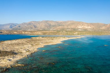 Yunanistan 'ın Girit kentindeki Elafonisi plajının geniş hava manzarasında genişleyen bir kumluk, sığ turkuaz su ve parlak, bulutsuz bir gökyüzü altında engebeli tepeler yer alıyor. Sahne güneşli, açık ve doğal olarak huzurlu..