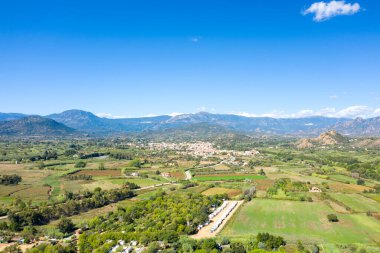 Lotzorai, Sardinya 'nın geniş hava manzarası, açık mavi bir gökyüzünün altında engebeli dağların dramatik bir zemin oluşturduğu yeşil tarlaların, dağınık ağaçların ve uzak bir köyün bir kısmını gösteriyor..