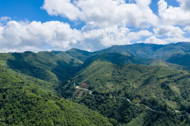 Kuzey Laos 'ta büyük, dalgalı bulutlarla dolu bir gökyüzünün altındaki sık ormanlık tepeleri kesen dolambaçlı bir yolu olan yeşil bir dağ vadisinin geniş manzarası. Manzara katmanlı yamaçlar, yemyeşil bitki örtüsü ve dramatik bir doğaya sahiptir.