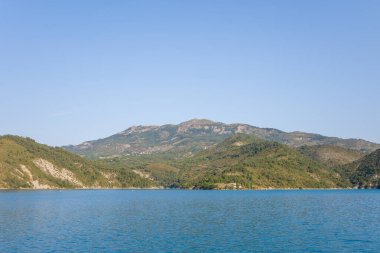 Castillon Gölü 'nün sakin mavi suları berrak, bulutsuz bir gökyüzünün altındaki yumuşak eğimli yeşil tepelere ve engebeli dağ tepelerine doğru uzanıyor..