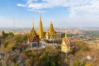 Süslü altın pagodalar ve tapınak yapılarından oluşan hava manzarası, kurak mevsim bitki örtüsü ve Kamboçya 'nın Battambang yakınlarındaki geniş tarım ovalarıyla çevrili Phnom Sampov' un kayalık zirvesinde tünemişti. Kompozisyon vurgulanıyor